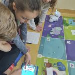 Troje dzieci siedzi na podłodze obok kolorowej maty i programują trasę robota na tablecie.