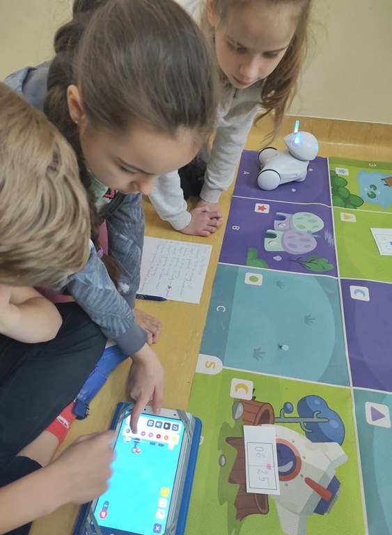 Troje dzieci siedzi na podłodze obok kolorowej maty i programują trasę robota na tablecie.