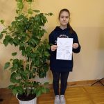 Dziewczynka ubrana na czarno stoi w sali, w rękach trzyma portret Piłsudskiego wykonany ołówkiem, obok niej duży kwiat doniczkowy