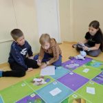 Dwóch chłopców siedzi na podłodze obok kolorowej maty i rozwiązuje zadania na kartce, podczas gdy dziewczynka programuje robota.