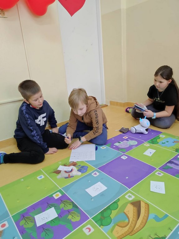 Dwóch chłopców siedzi na podłodze obok kolorowej maty i rozwiązuje zadania na kartce, podczas gdy dziewczynka programuje robota.