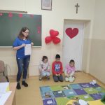 Troje siedzących na podłodze dzieci i jedna stojąca dorosła kobieta patrzą na leżącą na podłodze kolorową matę, na której stoi biały robot i rozłożone są białe karteczki.