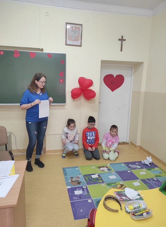 Troje siedzących na podłodze dzieci i jedna stojąca dorosła kobieta patrzą na leżącą na podłodze kolorową matę, na której stoi biały robot i rozłożone są białe karteczki.