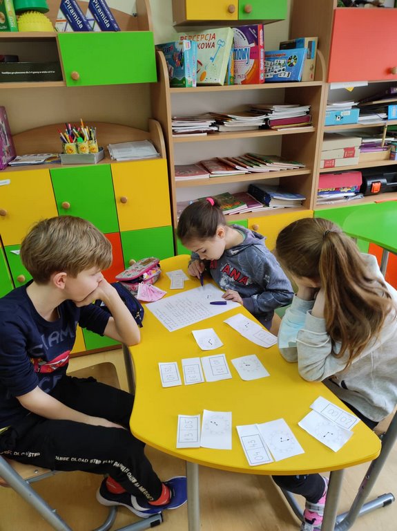 Dwie dziewczynki i jeden chłopiec siedzą przy żółtym stoliku i rozwiązują zadanie na kartce.