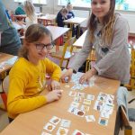 Dwie uśmiechnięte dziewczynki rozkładają na ławce obrazki z produktami. W tle inne pracujące grupy.