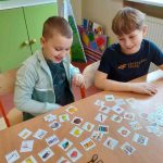 Dwóch uśmiechniętych chłopców siedzi przy stoliku, na nim rozłożone obrazki.