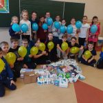 20 dzieci trzyma balony w kolorach niebieskich i żółtych. Przed nimi na podłodze materiały opatrunkowe, higieniczne.