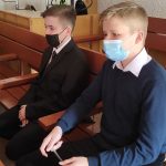 Chłopiec w ciemnym garniturze siedzi w brązowej, drewnianej ławce, oczekując na rozpoczecie konkursu. Obok niego drugi chłopiec, w granatowym swetrze i białej koszuli