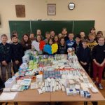 25 uczniów zgromadzonych wokół dużego stołu na którym leżą zgromadzone materiały opatrunkowe i higieniczne. Dwie dziewczyny trzymają w rękach duże serca: biało - czerwone i niebiesko - żółte.