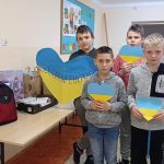 Pięciu chłopców trzyma w rękach niebiesko-żółte serca różnej wielkości. Na największym sercu napis: Solidarni z Ukrainą. Na stoliku obok chłopców papierowe torby i kartony z rzeczami dla uchodźców z Ukrainy oraz plecaki szkolne wypełnione zeszytami i przyborami szkolnymi.