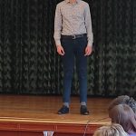 Szczupły chłopiec o ciemnych włosach, w jasnej koszuli, stoi na scenie. Przed nim stół jury, przykryty niebieskim obrusem. Z prawej strony widoczne głowy członków jury.