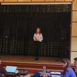 Na środku sceny, na tle ciemnozielonej kotary, stoi dziewczyna w czarnych spodniach i jasnej bluzce. Po lewej stronie niebieska tablica dekoracyjna. Na tablicy portret Józefa Piłsudskiego i otwarta książka. Po lewej stronie, przy stole siedzą członkowie jury. Dwie kobiety i jeden mężczyzna.