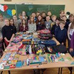 grupa uczniów zgromadzonych wokół dużego stołu na którym leżą zgromadzone materiały piśmiennicze, biurowe, przybory, plecaki i piórniki szkolne.