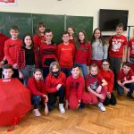 Grupa uczniów ubranych na czerwono stoi w sali lekcyjnej. Jeden z uczniów trzyma rozłożony, czerwony parasol