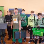 8 uczniów ubranych na zielono stoi w sali lekcyjnej. W rękach trzymają koszyki, walizki, wiaderka