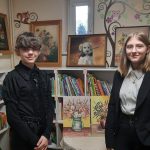 Dwie uczennice, ubrane na ciemno, stoją na tle prezentowanych obrazów.