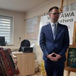 Malarz trzyma akordeon, obok stoi wójt gminy Tomasz Banek. Scena we wnętrzu biblioteki