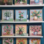 Na półkach regału ułożone są obrazy z kwiatami