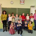 Jedenaścioro dzieci i dwie kobiety stoją na tle tablicy, trzymając dyplomy i czekolady zdobyte w konkursie.