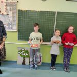 Dwie uśmiechnięte dziewczynki, chłopiec i kobieta stoją w klasie na tle tablicy trzymając czekolad w dłoniach. Grupowe - Jedenaścioro dzieci i dwie kobiety stoją na tle tablicy, trzymając dyplomy i czekolady zdobyte w konkursie.