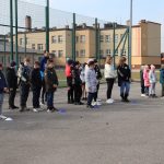 Uczniowie stoją na asfaltowym boisku. W tle budynek szkoły