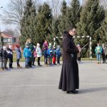 Ksiądz stoi na asfaltowym boisku, w ręku trzyma kartkę, z której odczytuje modlitwę, w tle uczniowie i nauczyciele