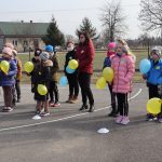 Grupa dzieci i nauczycielka na boisku szkolnym trzyma w rękach żółte i niebieskie balony