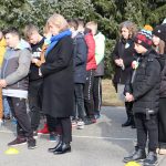 Kobieta w czarnym płaszczu stoi obok grupy uczniów odmawiających modlitwę