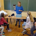 Kobieta w niebieskim swetrze trzyma w prawej ręce kubek, który ma dwa ucha. Przed nią, na stoliku, niebieskie pudełko z zawartością gry.