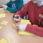 Dwaj chłopcy, jeden w czerwonej bluzie, drugi w zielonej, wycinają kwiaty z żółtego papieru.