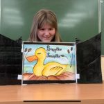 Uśmiechnięta dziewczynka prezentuje opowieść w teatrzyku kamishibai. Na ilustracji teatrzyku żłółta kaczka