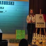 Dziewczynka w białej bluzce i czarnych spodniach stoi na scenie przed mikrofonem i recytuje wiersz