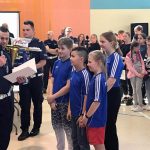 Grupa osób, policjant odczytuje wynik zawodów, przed nim soi czworo uczniów w niebieskich koszulkach