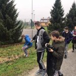 Grupa uczniów, jeden chłopiec trzyma w rękach worek, drugi wskazuje palcem na coś, co trzeci chłopiec podnosi z trawy