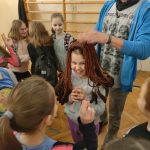 Uśmiechnięta dziewczynka zakłada bordową gliniana perukę.