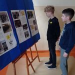 Dwóch chłopców stoi przed tablicą, na której znajdują się informacje na temat powstania w getcie warszawskim. Na tablicy i na bluzach chłopców widoczne żółte żonkile.