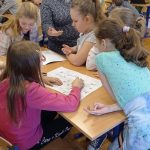 Kobieta w szarej sukience tłumaczy dziewczynkom zasady gry