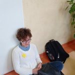 Dziewczynka o ciemnych włosach i w białym sweterku siedzi na tle białych drzwi. Do sweterka ma przypięty żółty żonkil.