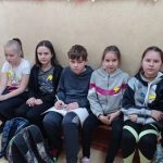 Pod ścianą, na drewnianej ławce siedzą cztery dziewczynki i jeden chłopiec. Na ubraniach dziewczynek znajdują się żółte żonkile.