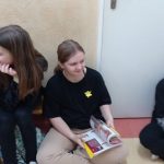 Przed drzwiami do klasy siedzi dziewczynka w czarnej bluzce. Na bluzce przypięty żółty żonkil.