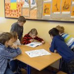 Dziewczynka w czerwonym sweterku czyta instrukcję gry, kilku uczniów przysłuchuje się z zainteresowaniem.