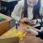 Dziewczynka o długich ciemnych włosach skleja elementy żonkila. Przed nią, w kartonowym pudełku kilkadziesiąt gotowych kwiatów.