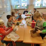 Dzieci siedzą przy stolikach i wsypują ziemię do doniczek.