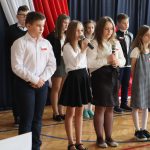 Grupa uczniów na sali gimnastycznej. Dwie dziewczynki trzymają mikrofon, jedna z nich recytuje wiersz.