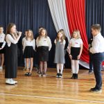 Dziewczynka i chłopiec rozmawiają ze sobą przez mikrofon. W tle uczniowie w strojach galowych.