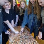 pięć uśmiechniętych dziewczyn przyglada się tablicy wyklejonej zdjęciami głów ludzkich. Obok nich stoi jeden zamyślony chłopiec.