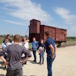 Uczniowie stoją na tle drewnianego wagonu towarowego, którym przywożono do obozu więźniów. W tle ogrodzenie z drutu kolczastego.