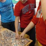 Trzech chłopców stoi nad tablicą wyklejoną zdjęciami głów kobiet i mężczyzn, wskazują palcami podobiznę Miłkowskiego