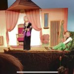 Na scenie kobieta w czarnej sukni rozmawia z mężczyzną siedzącym na podłodze