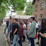 Uczniowie, w słuchawkach na uszach, stoją na jednej z obozowych uliczek.W tle piętrowy budynek z czerwonej cegły.